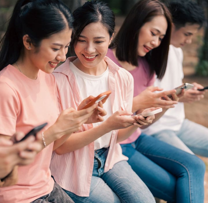 Four young people sit closely together on a bench outdoors, smiling and looking at their smartphones. Dressed in light, casual clothes, they appear engaged—possibly sharing or viewing something on their devices together. The softly blurred background hints at a vibrant setting typical of many Asia Pacific cities.