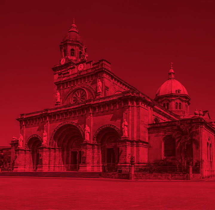 A red-tinted photo of a grand cathedral with arched entrances, ornate columns, statues, and a clock tower on the left captures an architectural gem reminiscent of landmarks found throughout the Asia Pacific region. A large dome rises behind the building while the clear sky and bright daylight cast sharp shadows across an empty plaza in the foreground.