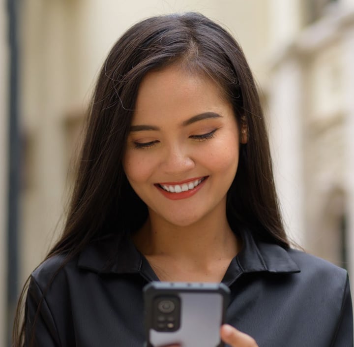 A woman with long dark hair and a black shirt smiles softly while looking down at a smartphone, standing outdoors in an Asia Pacific city with blurred buildings in the background.