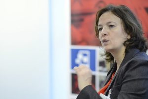 A woman with brown hair, wearing a dark blazer and orange top, gestures with her hand while speaking. The background is blurred, showing a wall with red and blue panels and partial symbols. She appears engaged and focused.