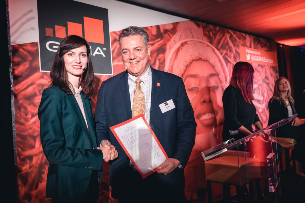 A woman in a green suit and a man in a gray suit shake hands and smile at a formal event. The man holds a document. Behind them is a red-themed backdrop with the GEIA logo, social media info, and a large smiling face. Other people stand near a podium.
