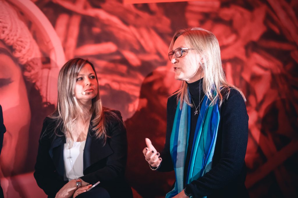 Two women sit and talk onstage against a red-toned background. The woman on the right gestures with her hand while speaking, wearing glasses, a blue scarf, and a black top. The woman on the left listens attentively, holding a phone or notepad in her lap.