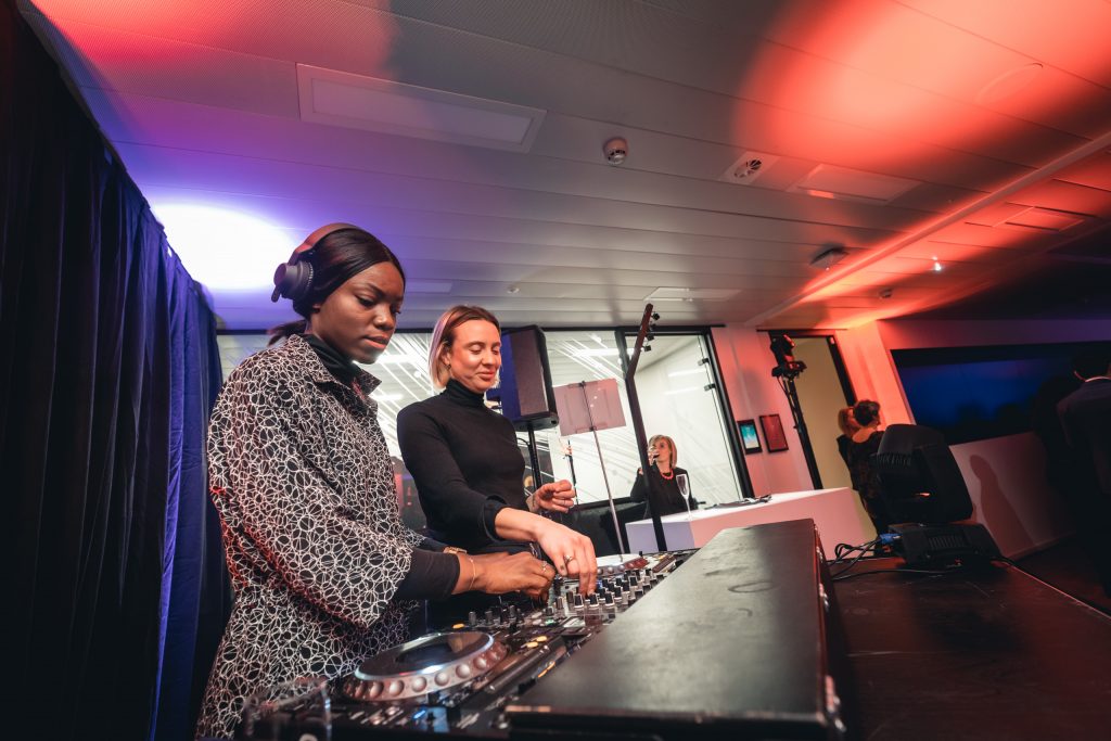 Two DJs stand behind a mixing console in a modern indoor venue with colored lighting. The DJ on the left, wearing headphones and a patterned jacket, adjusts controls, while the other, in a black turtleneck, smiles and operates the equipment. People and screens are in the background.