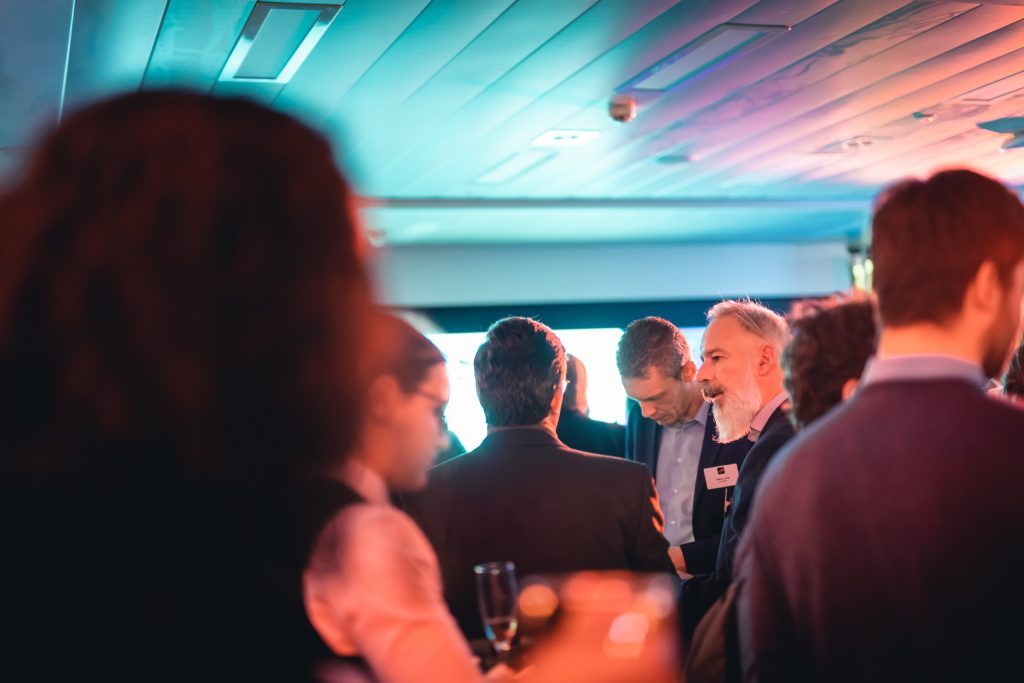 A group of people in semi-formal attire converse at an indoor networking event. The scene is softly lit with blue and pink hues. Several individuals are blurred in the foreground, while some are clearly visible talking in the background.