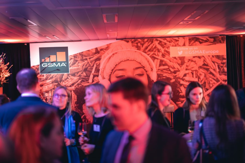 A group of people socialize at an indoor event, with a large GSMA banner in the background. The banner features a close-up of a woman's face in a knit hat. Warm, pink and orange lighting fills the room, creating a lively and inviting atmosphere.