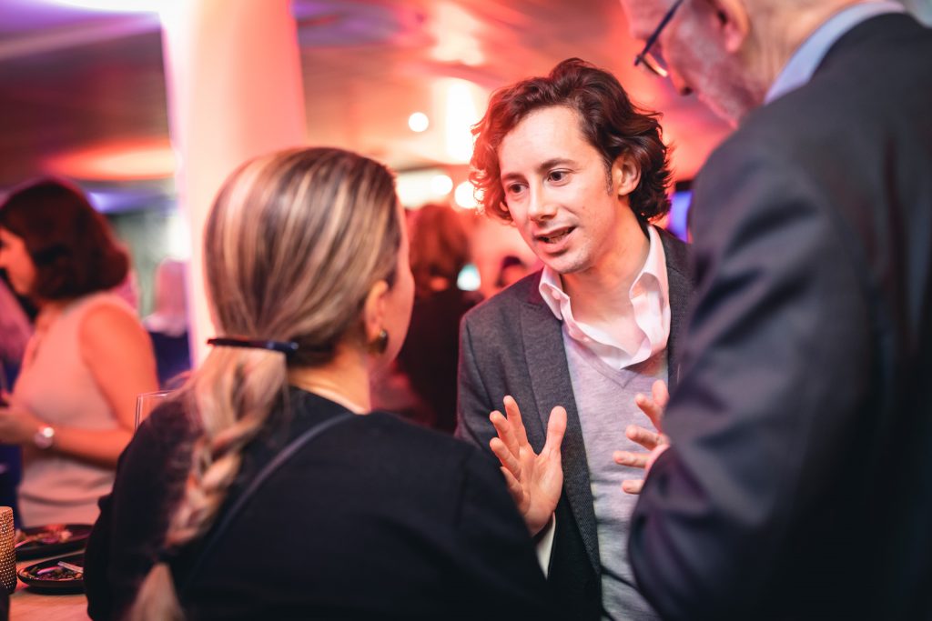 A man in a suit gestures while speaking to a woman with a braid at a lively indoor event. Another man stands beside them. The background is softly blurred, with people mingling and colorful lights creating a warm, vibrant atmosphere.