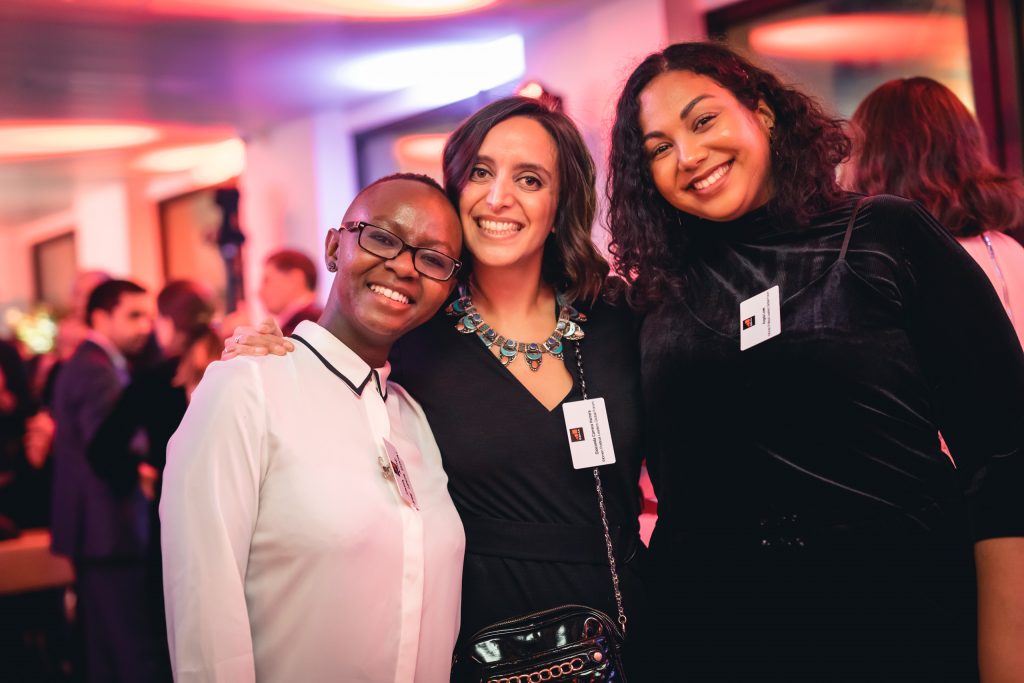 Three women stand close together, smiling at a formal indoor event. They wear name tags and dress in elegant, dark clothing. The background shows softly blurred people and warm pink lighting, creating a festive, welcoming atmosphere.