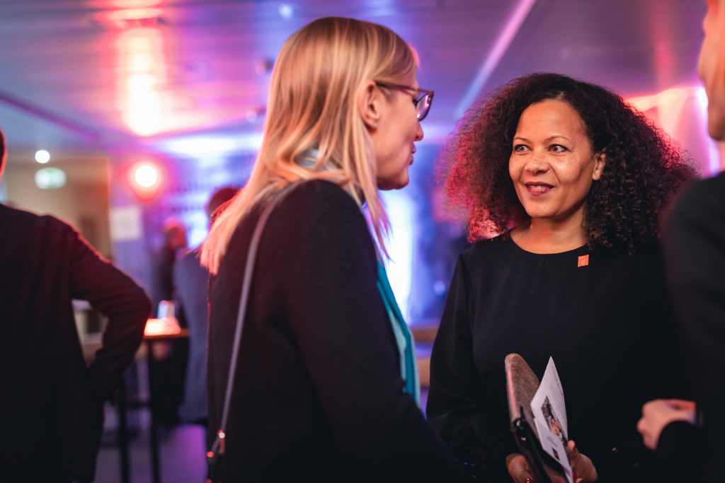 Two women, one with curly hair holding a paper and the other with straight blonde hair and glasses, are engaged in conversation at an indoor event with colorful lights and other attendees visible in the blurred background.