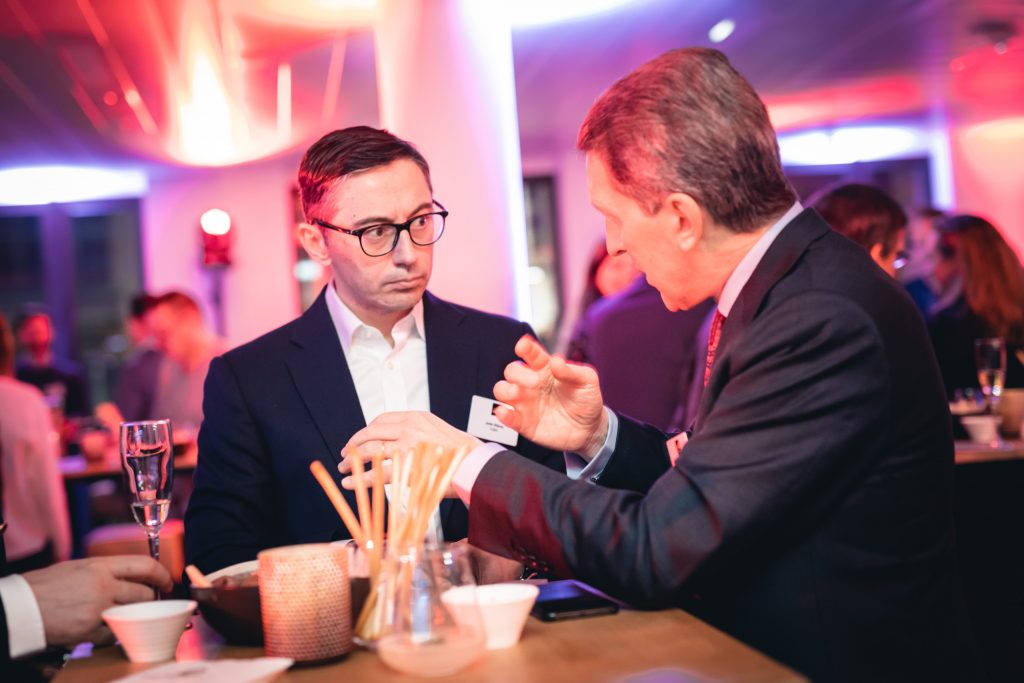 Two men in suits talk seriously at a dimly lit event, standing at a high table with drinks, breadsticks, and snacks. The background is blurred with other people and colorful lights, creating a lively atmosphere. One man gestures as he speaks.