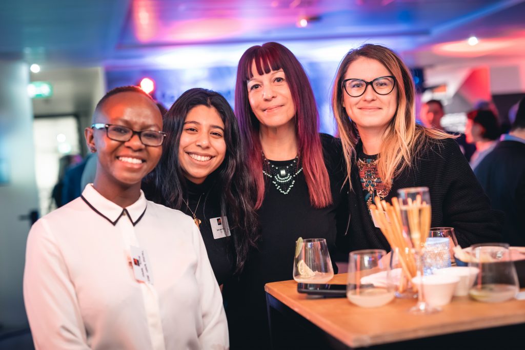 Four women smile together at an indoor social event, standing by a high table with drinks and snacks. The background is softly lit with blue and pink lights, and people can be seen mingling in the blurry distance.