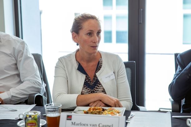 A woman with light brown hair tied back, wearing a white textured jacket over a navy patterned blouse, sits at a conference table with food, a drink, papers, and a nameplate reading “Marloes van Caspel.” She appears attentive, with a window in the background.