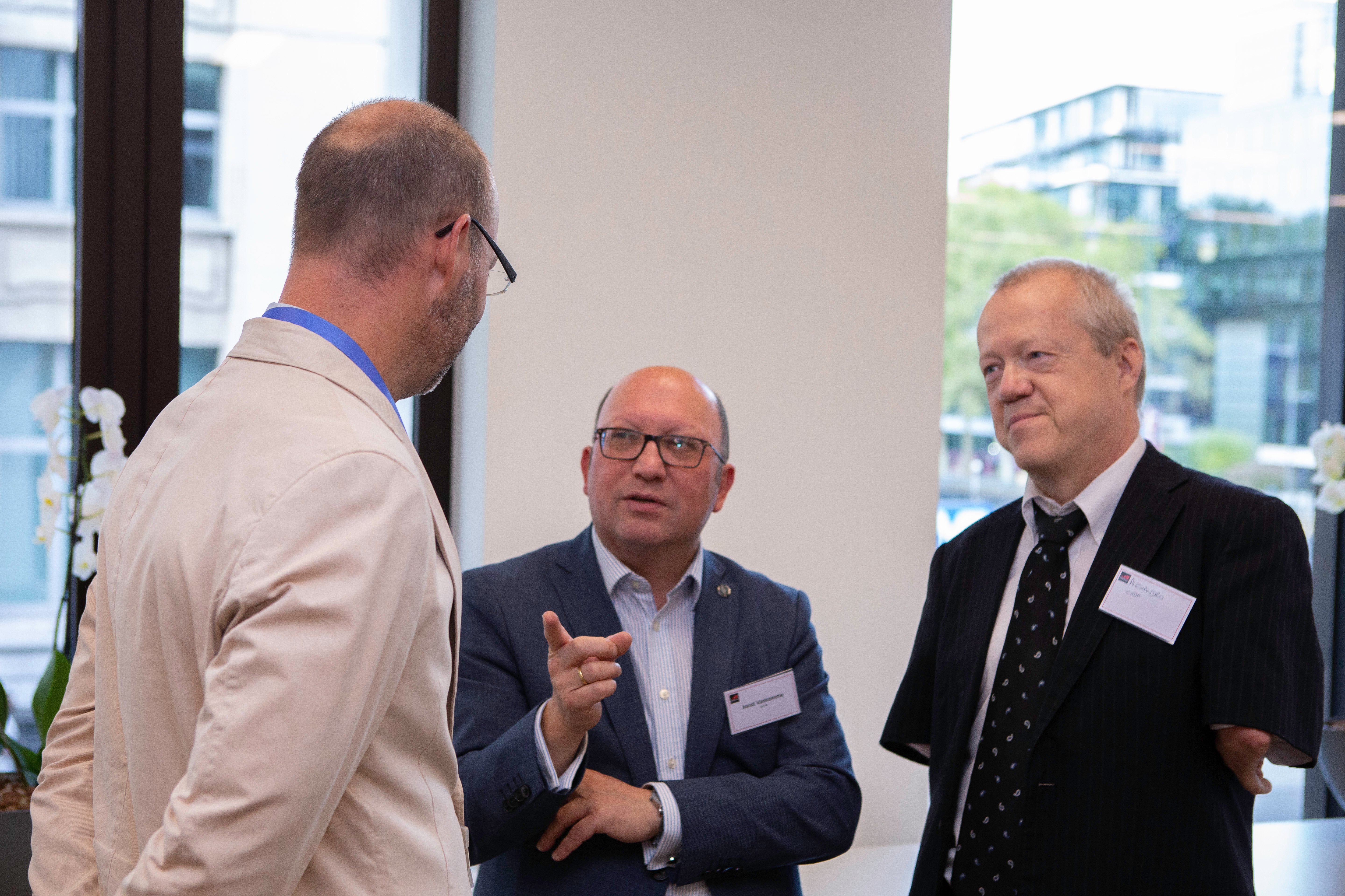 Three middle-aged men in business attire stand indoors discussing connected vehicles. One gestures while speaking, facing a man in a beige jacket. Large windows reveal modern buildings and hint at the dynamic world of connected cars outside.