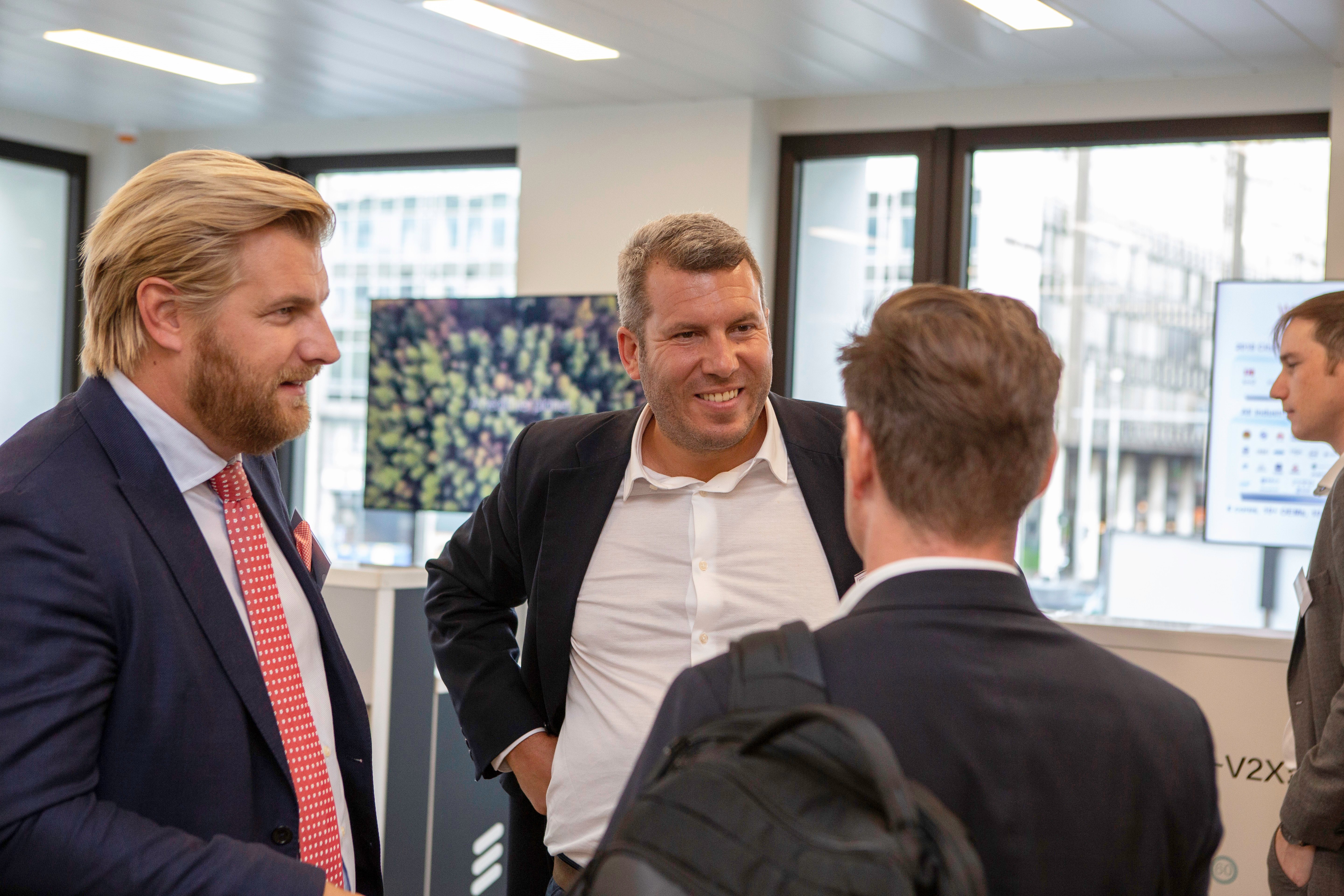 Three men in business attire discuss connected vehicles indoors near large windows. One, blonde with a beard, wears a navy suit and red tie. Another, gray-haired and smiling, stands with hands on hips. A fourth person is blurred in the background.