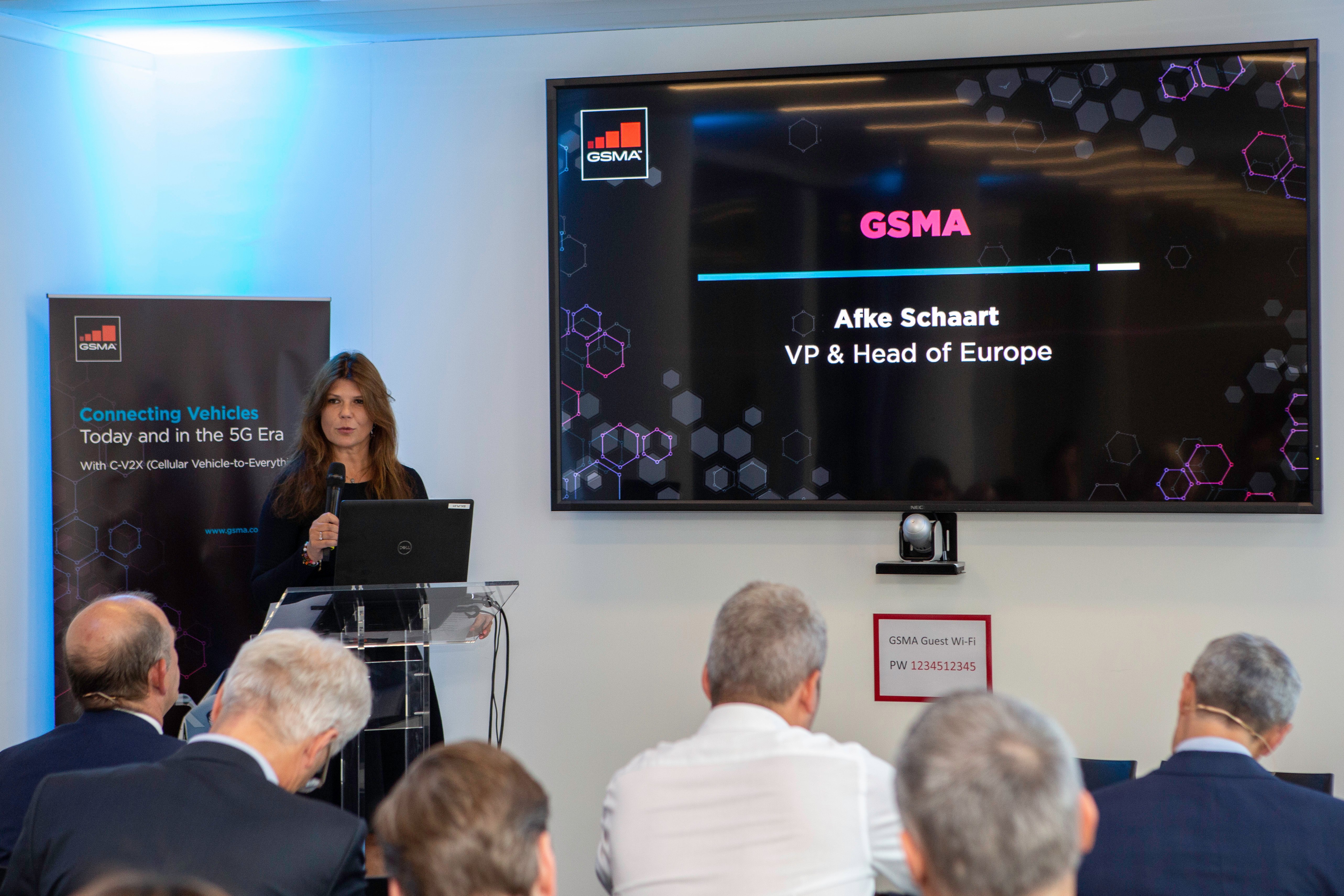 A woman stands at a podium addressing an audience. Behind her, a large screen displays: “GSMA, Afke Schaart, VP & Head of Europe.” Attendees listen as a banner on the left highlights connected vehicles and 5G technology.