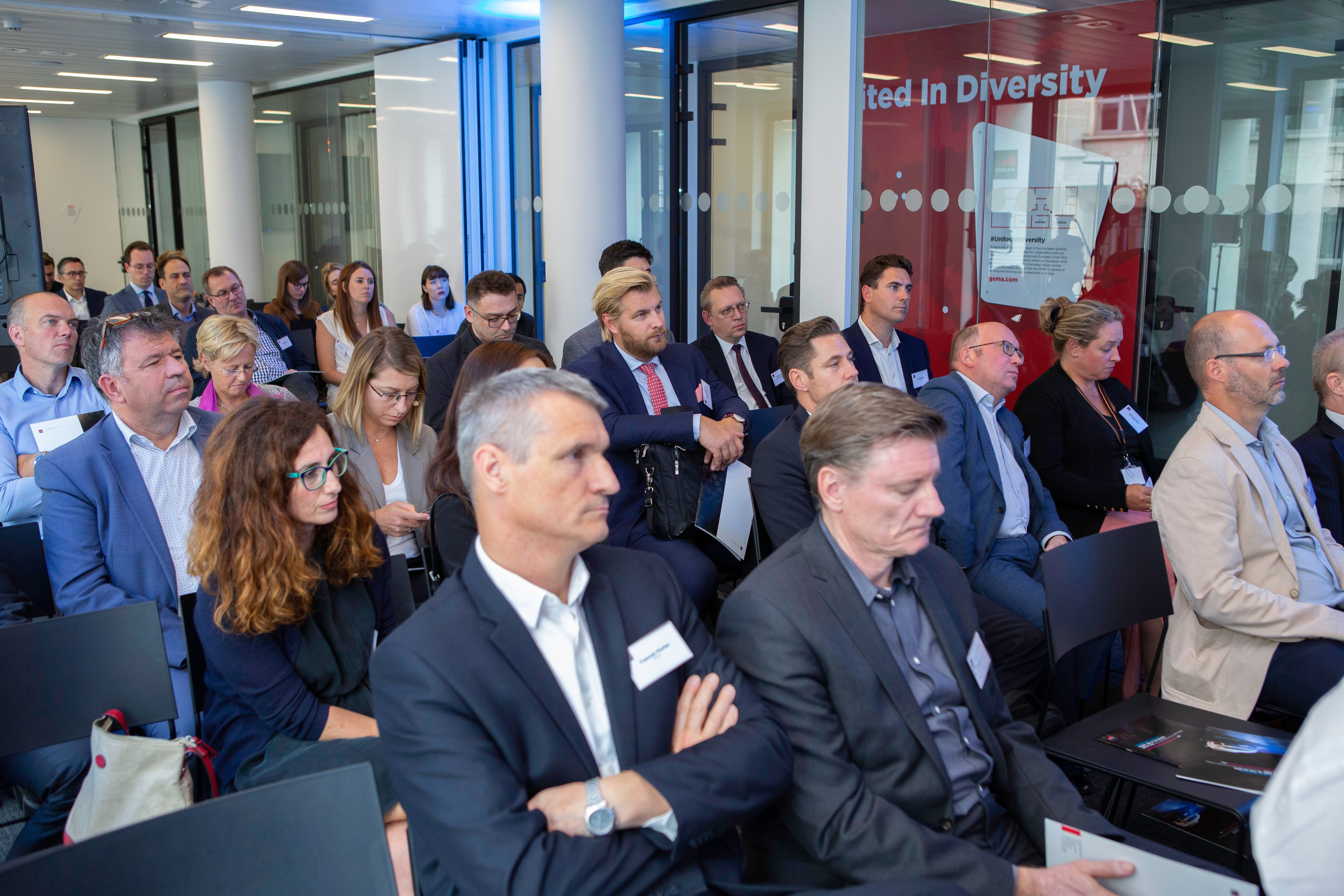A group of professionally dressed people sit attentively in a modern conference room, engaged in a discussion about connected vehicles. Glass walls and a red poster with “Diversity” are visible, adding to the formal, businesslike atmosphere.