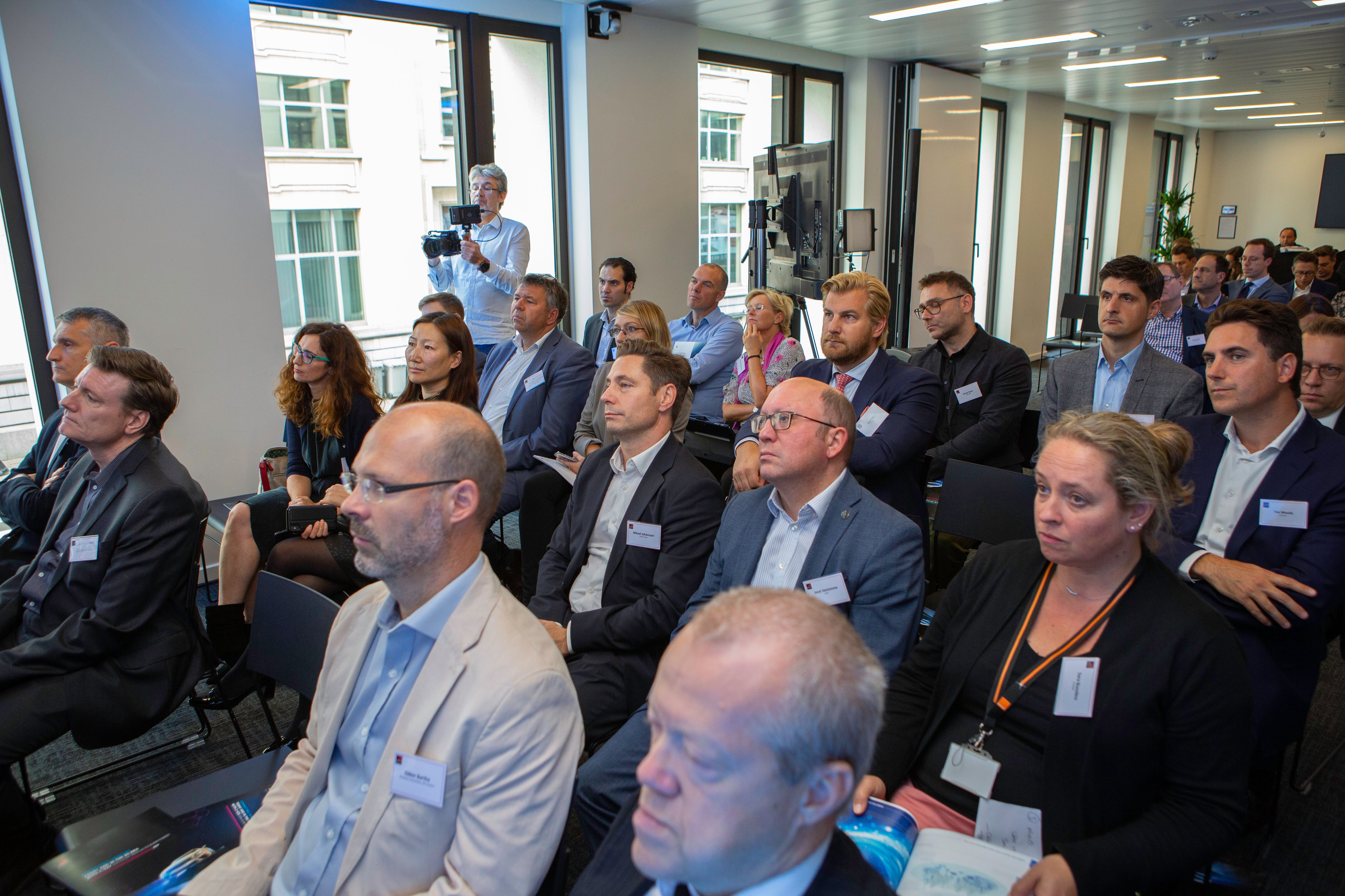 A group of professionally dressed adults sit attentively in rows at a business conference on connected vehicles in a modern, well-lit room. Some take notes; a photographer stands at the back. Large windows reveal another building outside. Name badges are visible on attendees.