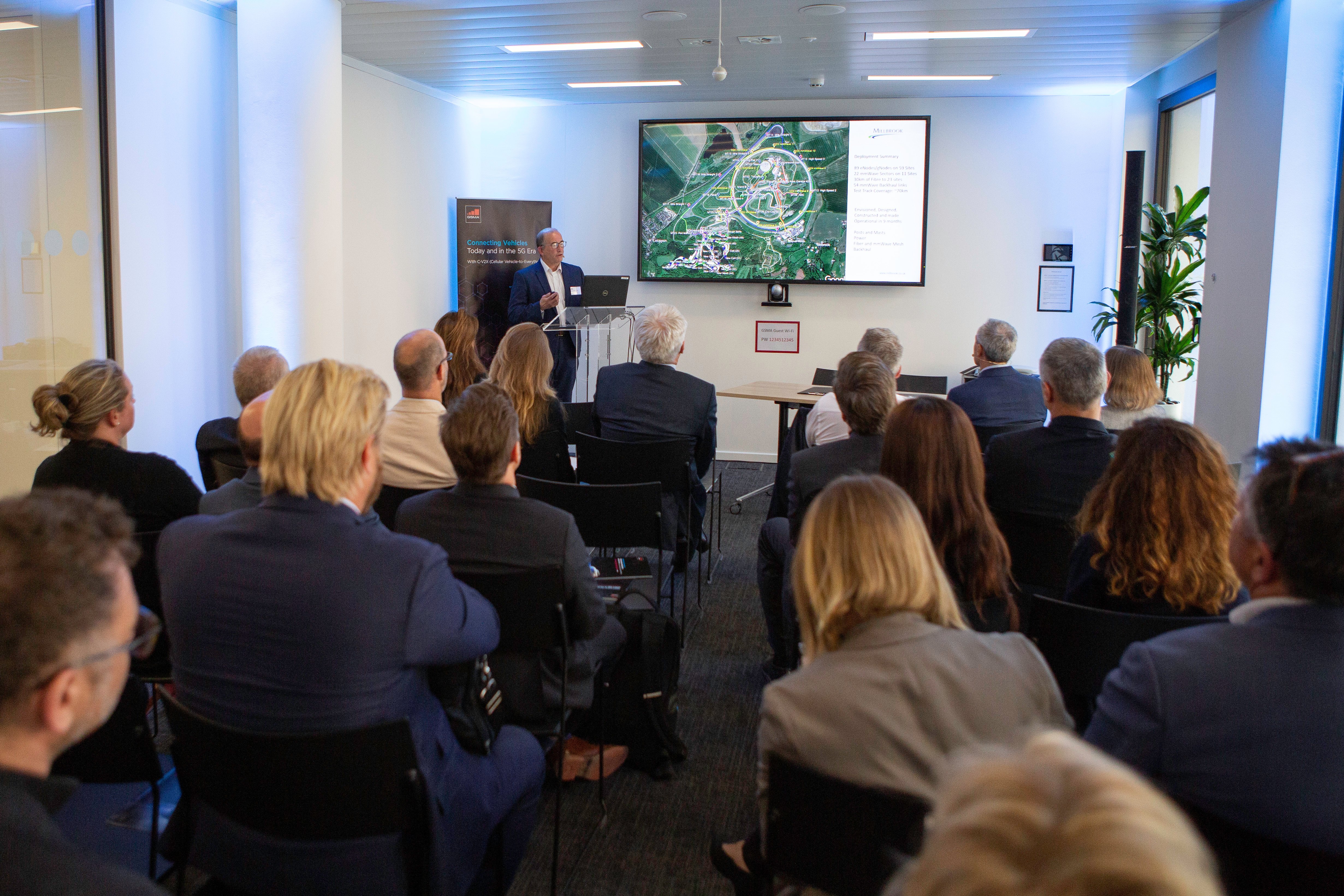 A group of people in business attire sit facing a presenter at the front of a conference room. The presenter stands by a podium and screen displaying a map and text about connected vehicles. The well-lit room features modern décor and plants on the right.
