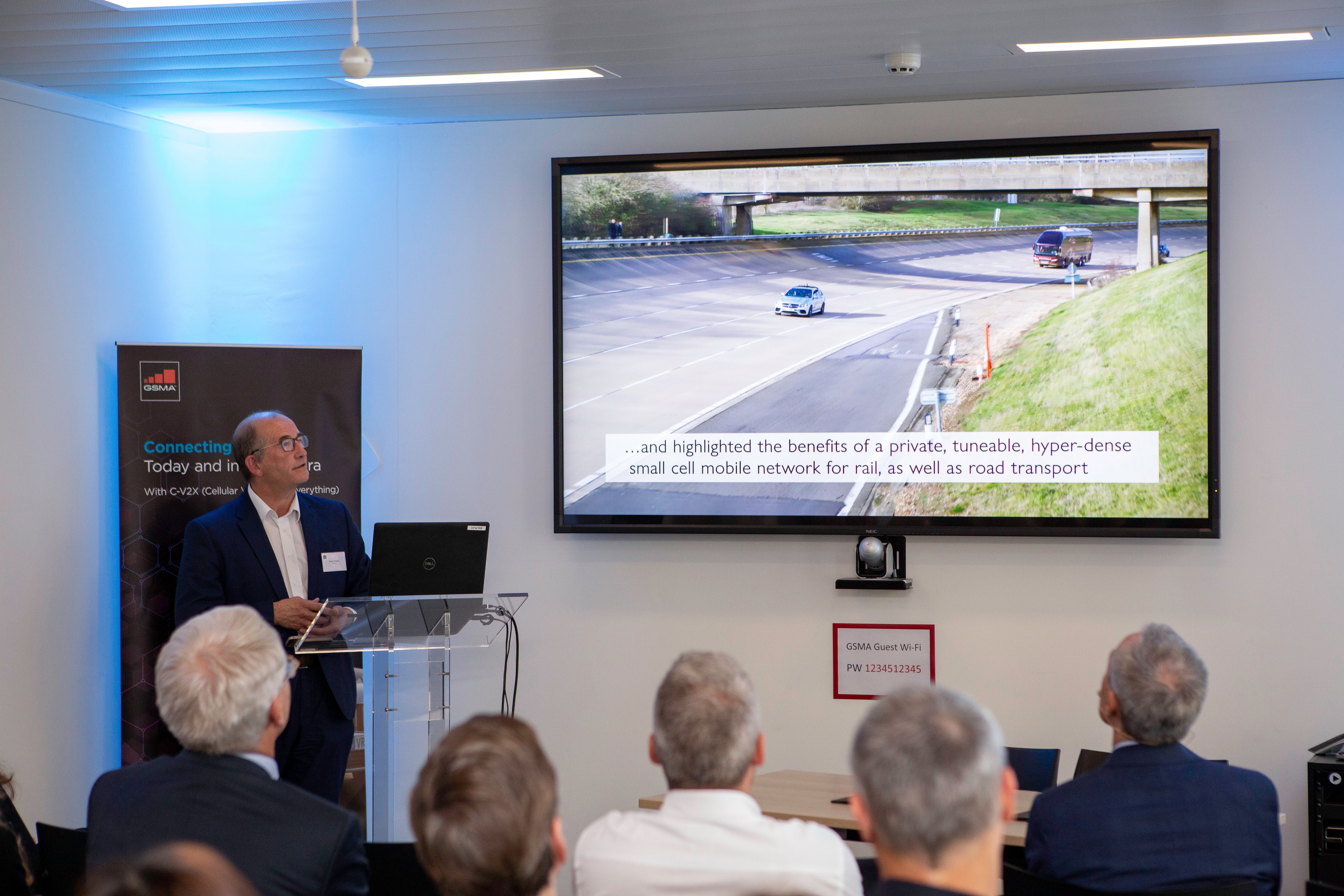 A man in a suit stands at a podium, presenting to a seated audience. A large screen shows connected vehicles on a highway and a caption about mobile networks for rail and road transport. A banner and emergency guide are visible in the modern room.