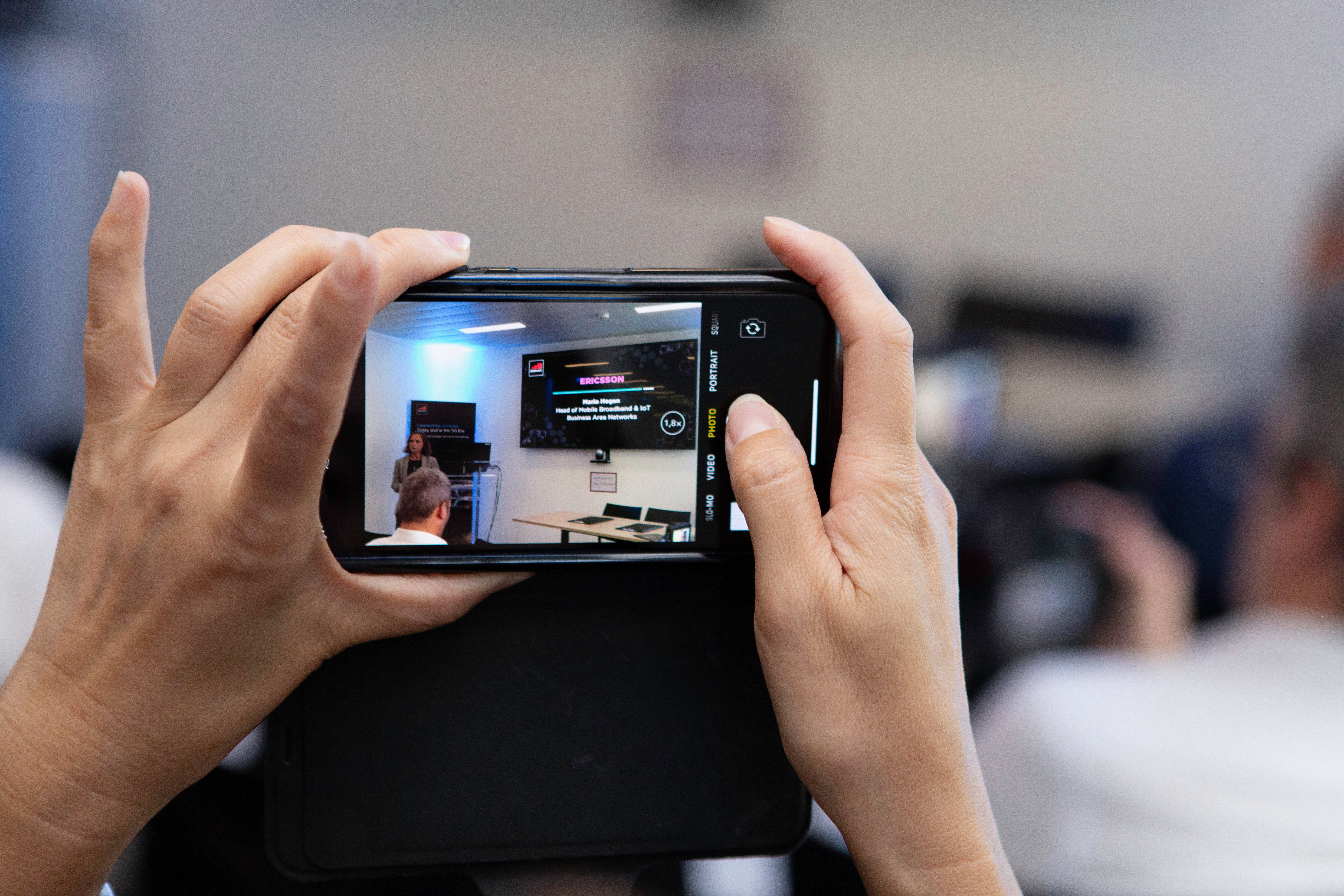 Close-up of hands holding a smartphone horizontally, capturing a presentation. On the screen, a speaker discusses connected vehicles in front of a projected slide; audience members and conference setting are blurred in the background.