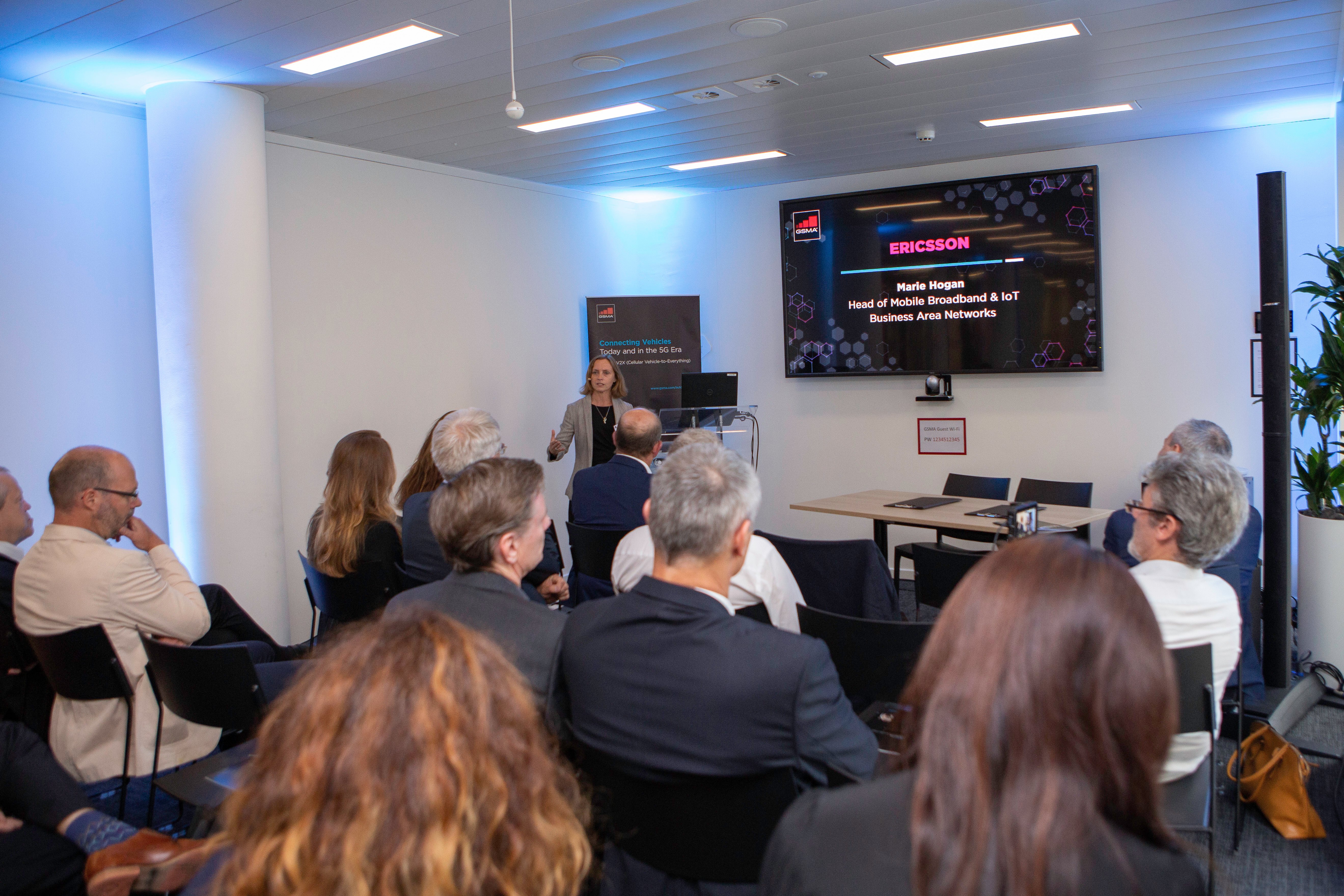A woman stands presenting in front of a seated audience in a modern conference room. A screen behind her displays the Ericsson logo and her name, “Helen Nagem, Head of Mobile Broadband & IoT, Business Area Networks,” discussing connected vehicles.