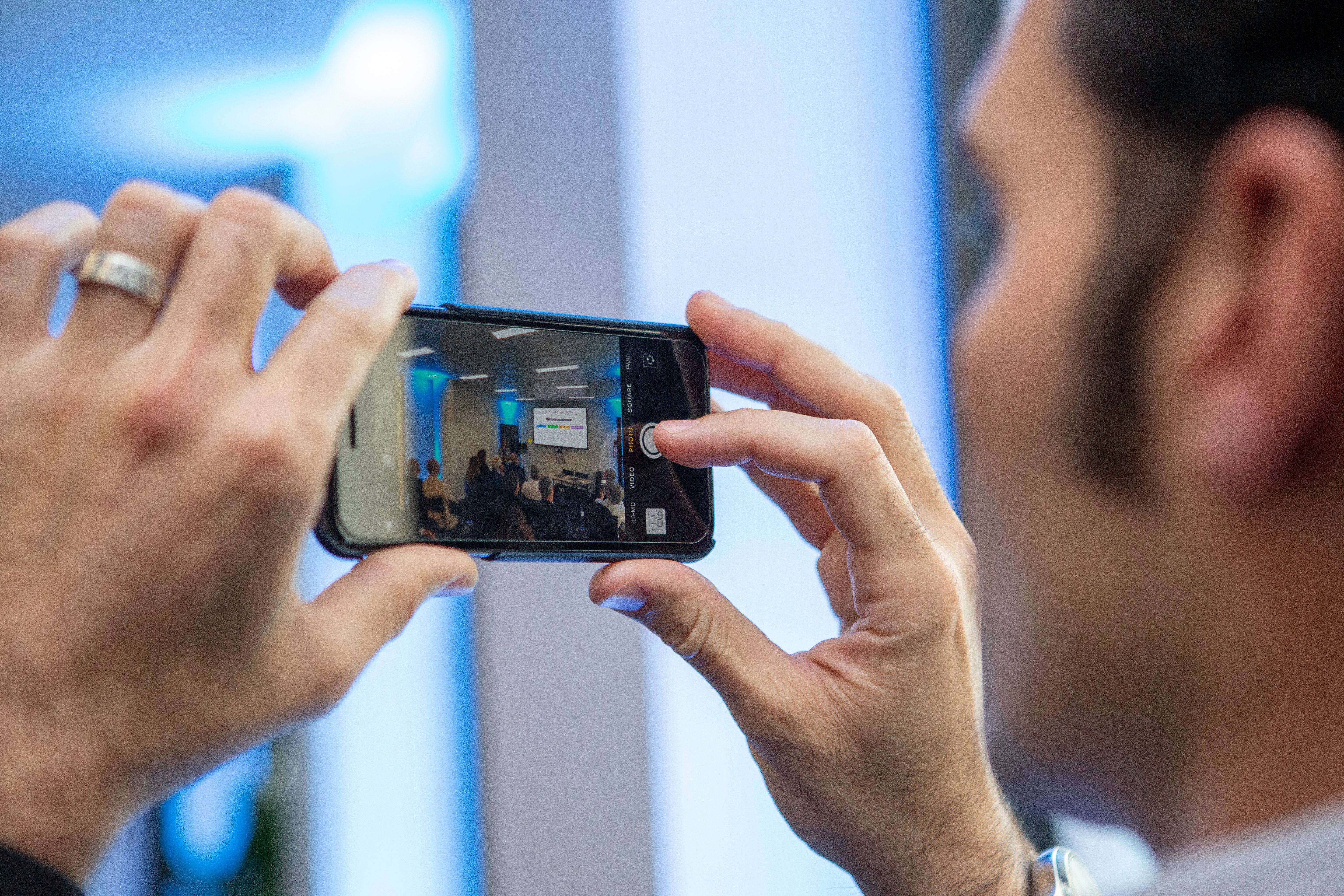 A person holds a smartphone horizontally, capturing a conference focused on connected vehicles. The phone screen shows an audience and a speaker in front of a projected slide in a brightly lit room with blue accents.