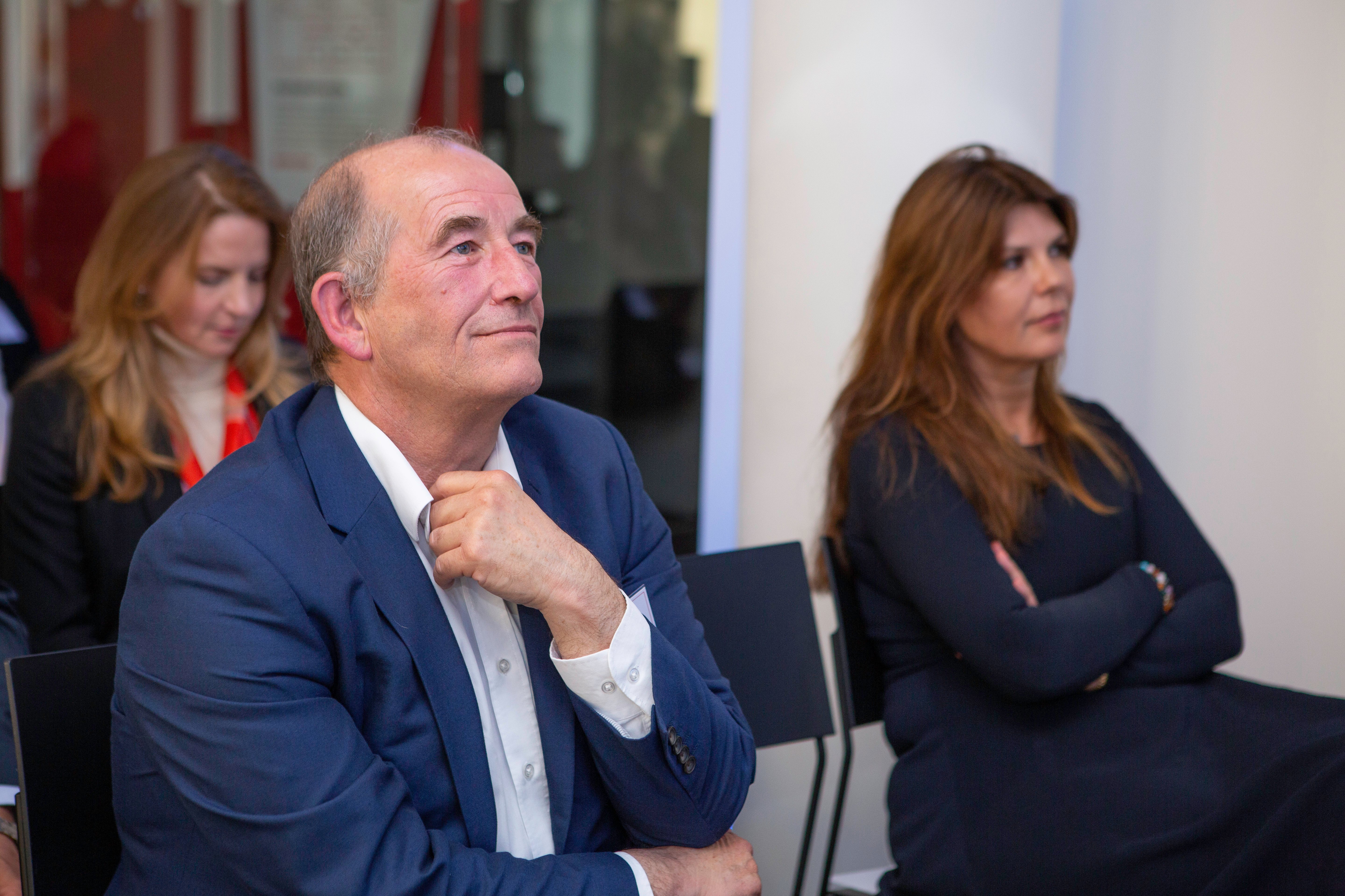 A man in a blue suit sits attentively with his hand on his chin, listening intently at an event focused on connected vehicles. Two women, one with long brown hair in black and another blonde, are seated in the background.