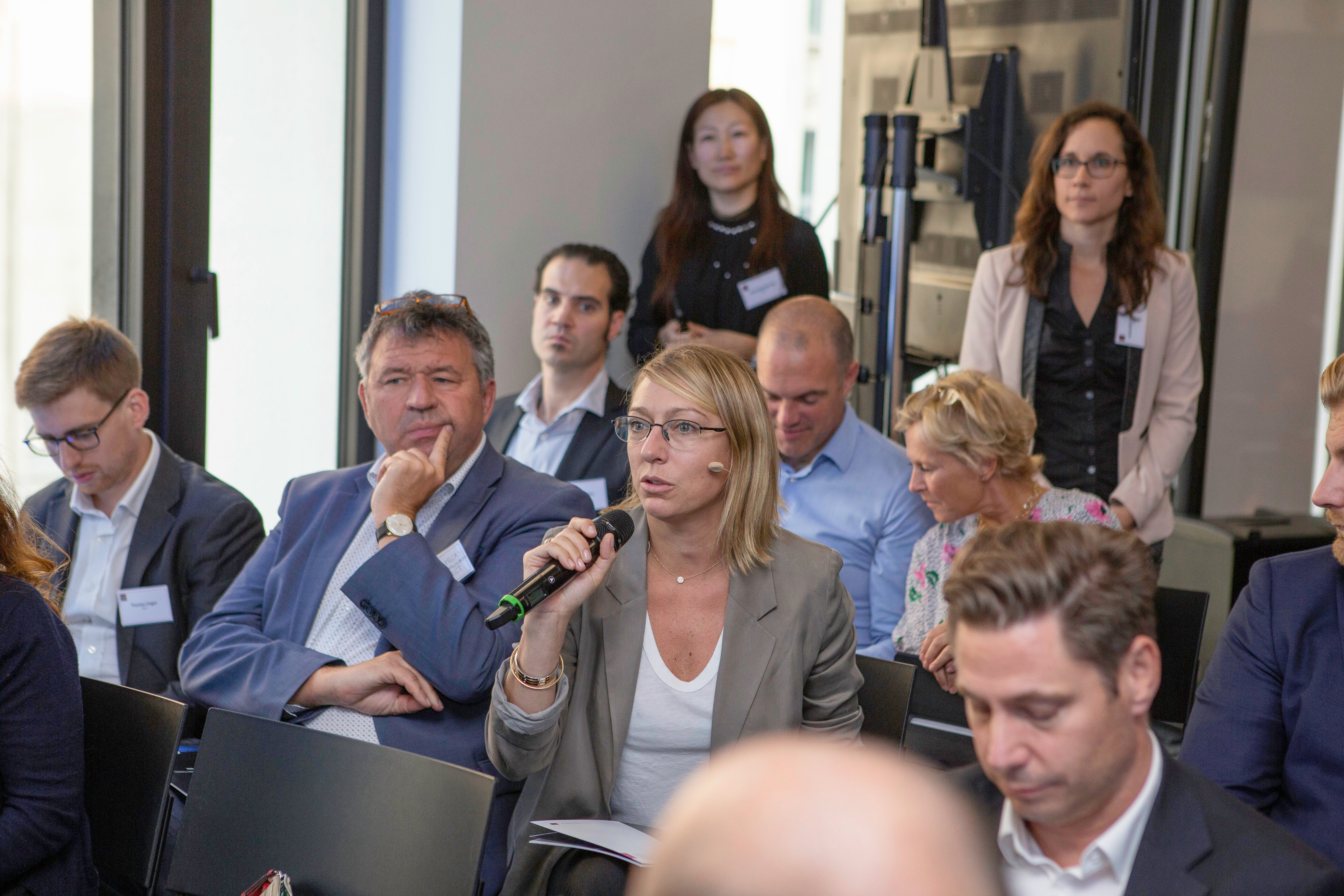 A woman holding a microphone speaks during a business event or conference on connected vehicles. She is surrounded by seated attendees in business attire, some listening attentively. A few people stand in the modern setting with large windows and neutral decor.