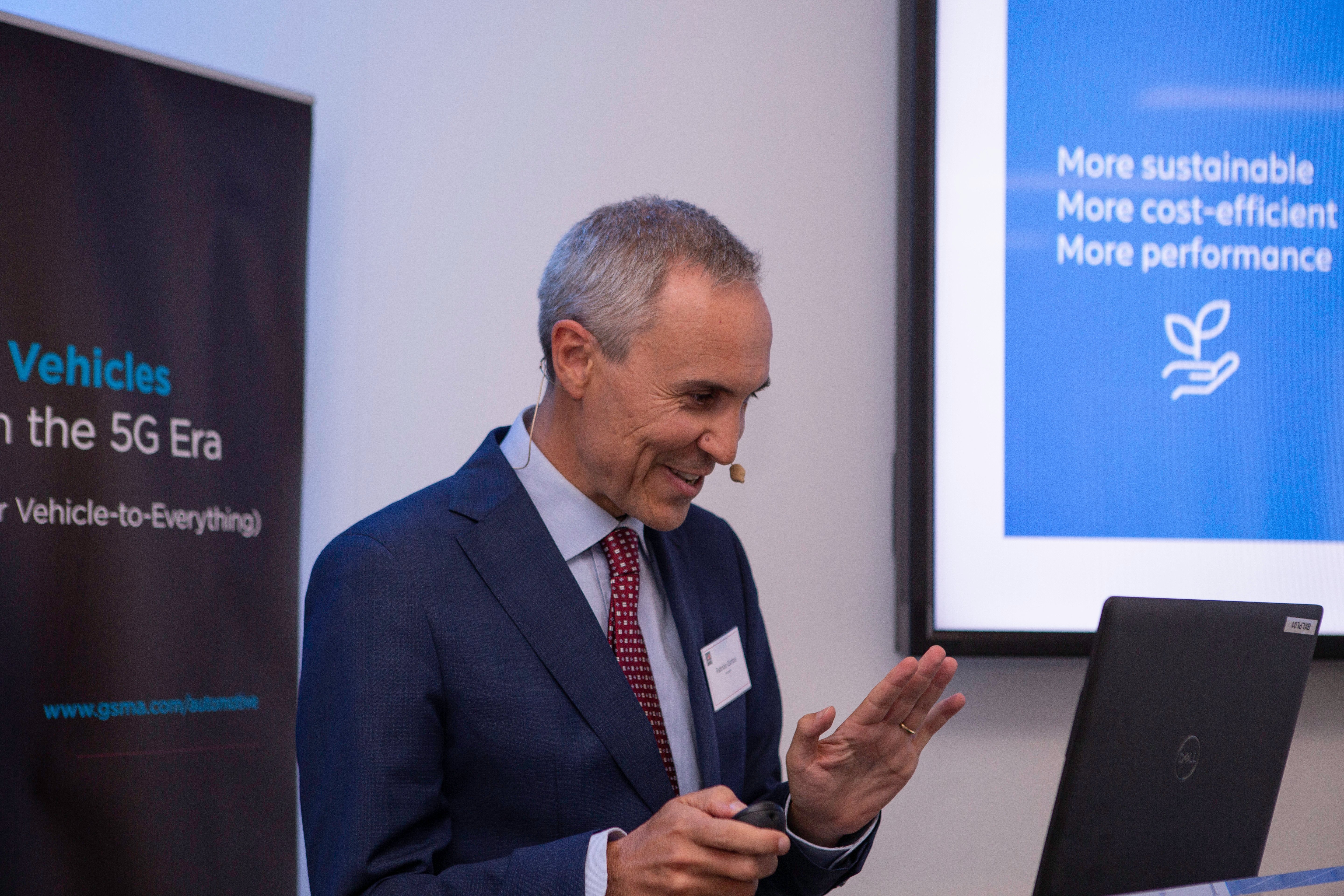 A man in a blue suit with a headset microphone gestures while speaking at a laptop. Behind him, screens display “More sustainable, More cost-efficient, More performance” with a leaf icon and reference connected vehicles and 5G cars.
