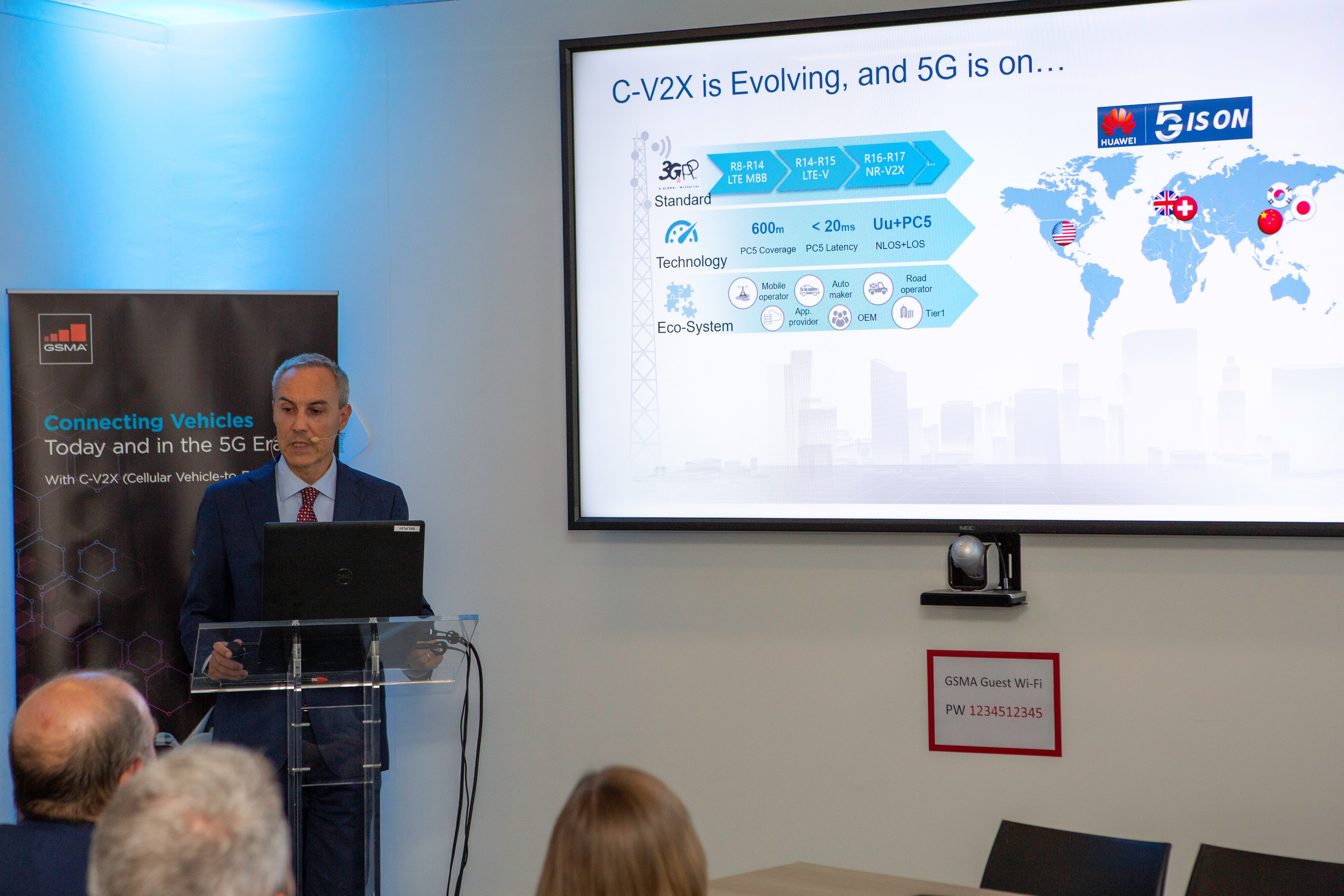 A man in a suit stands at a podium giving a presentation. Behind him, a large screen displays a slide about C-V2X, 5G, and Connected Cars, featuring a world map, charts, and icons. Audience members are seated near a GSMA banner on the left.