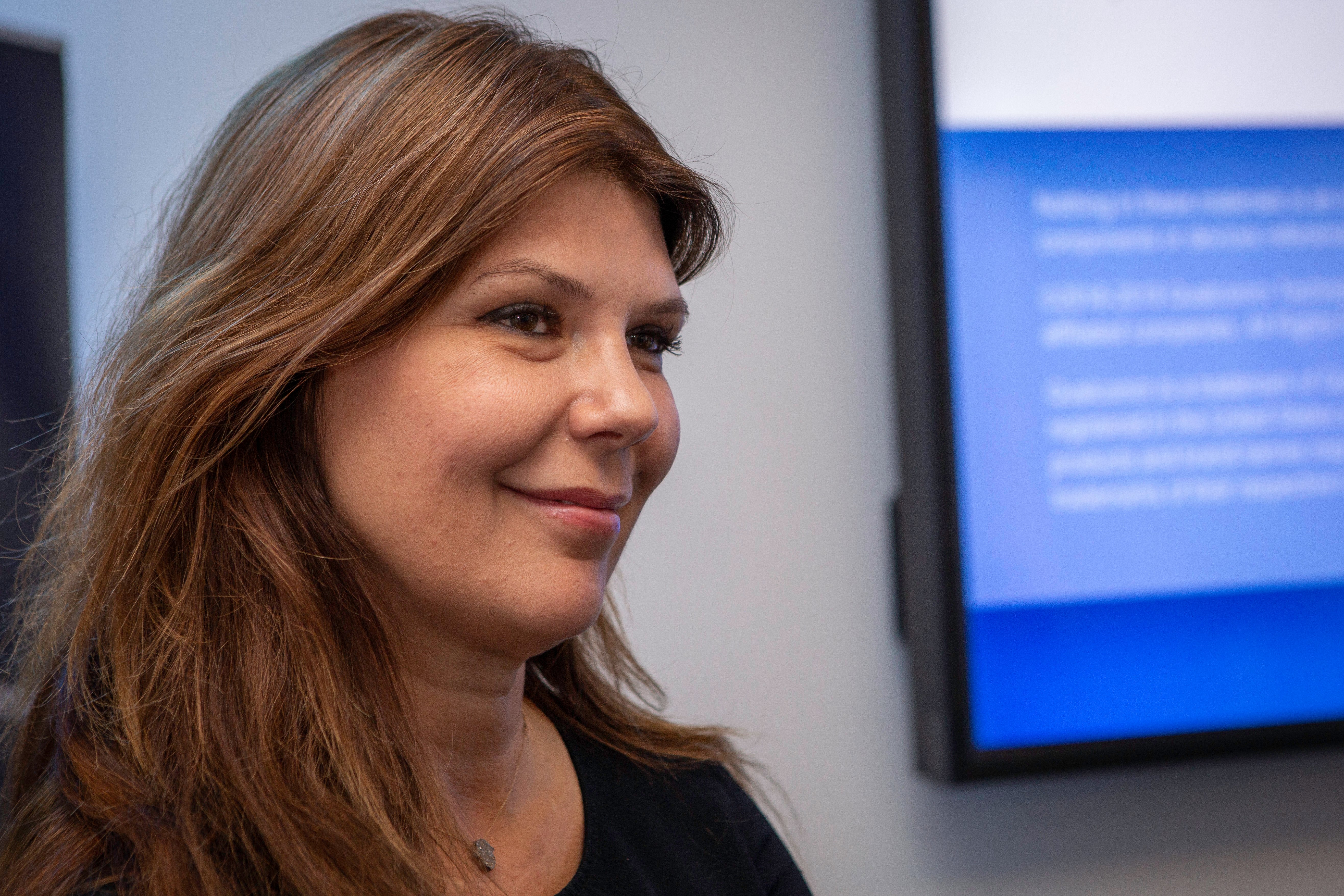 A woman with long, light brown hair smiles softly while looking to the left. She is indoors, wearing a dark top, and a blurred blue and white connected vehicles screen is visible in the background on the wall.