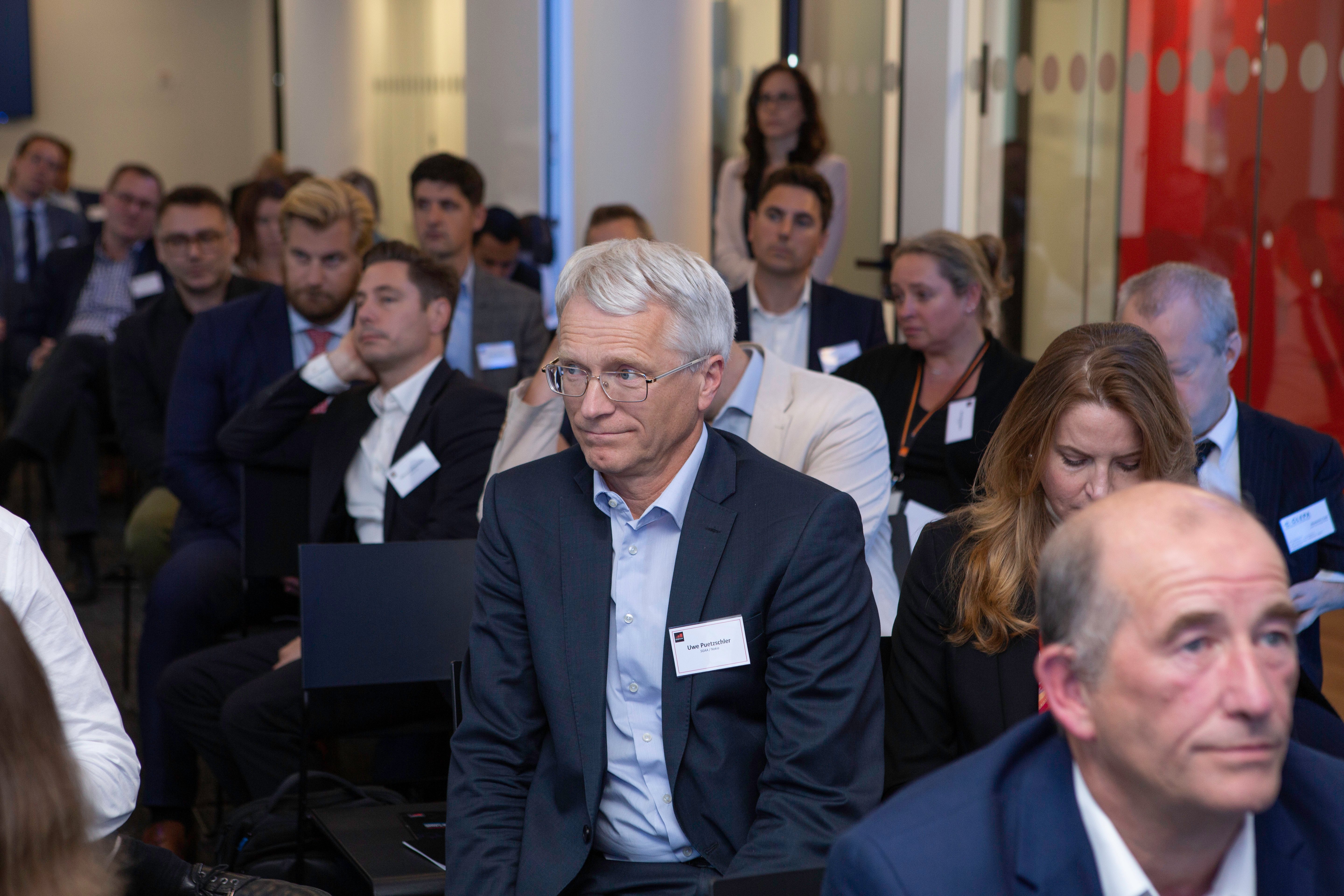 A group of people in business attire sit attentively in rows at a conference on connected vehicles. Name tags are visible, and the modern room with glass walls reflects the focus on innovations like connected cars.