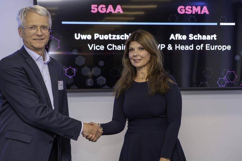 A man in a suit and glasses shakes hands with a woman in a dark dress. Behind them, a screen displays “5GAA,” “GSMA,” and their names and titles, highlighting connected vehicles at this professional event setting.