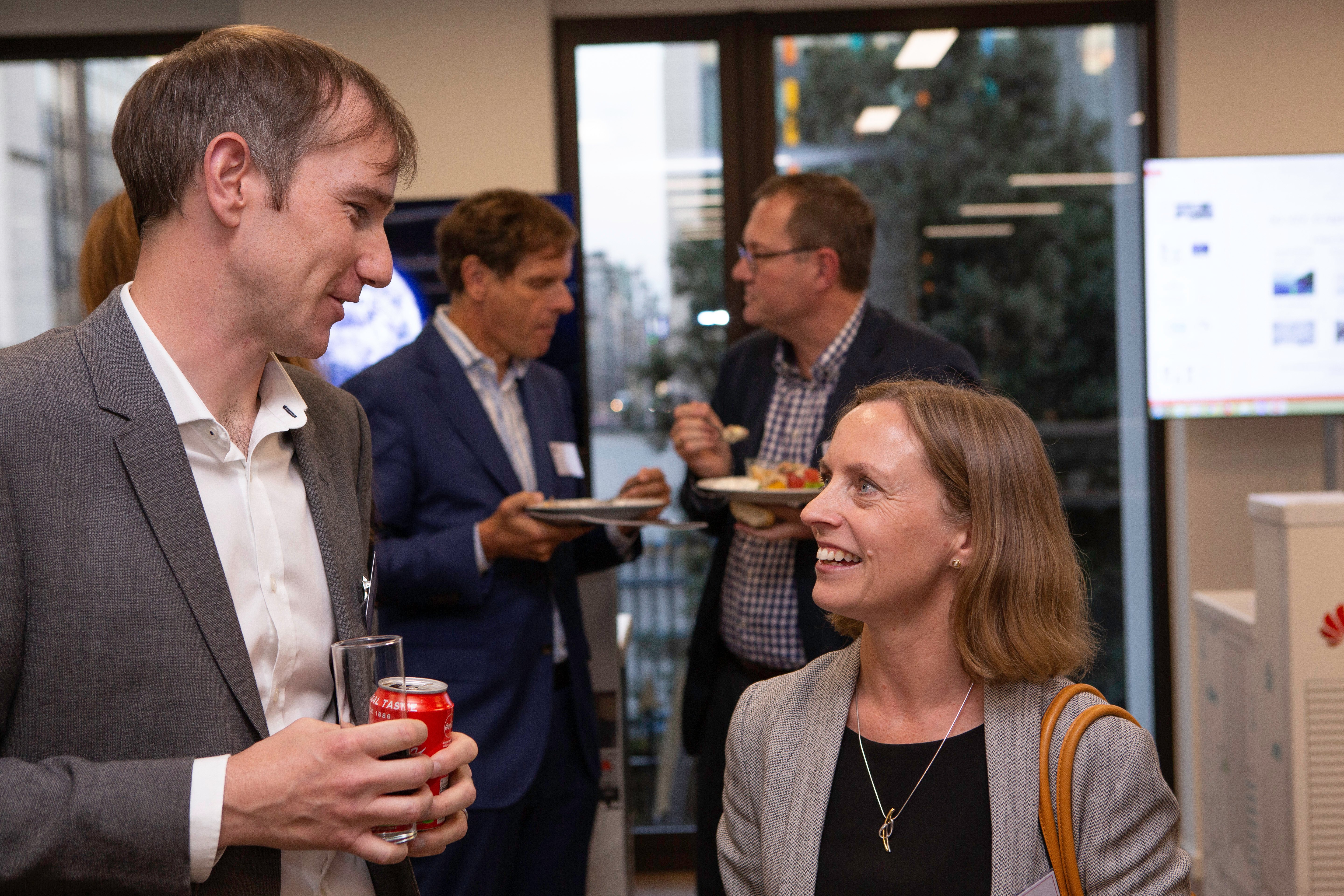 Two professionally dressed people stand and chat indoors; the man holds a glass and a soda can, while the woman smiles at him. In the background, two men in suits discuss connected cars as they eat. Large windows reveal trees and city buildings outside.