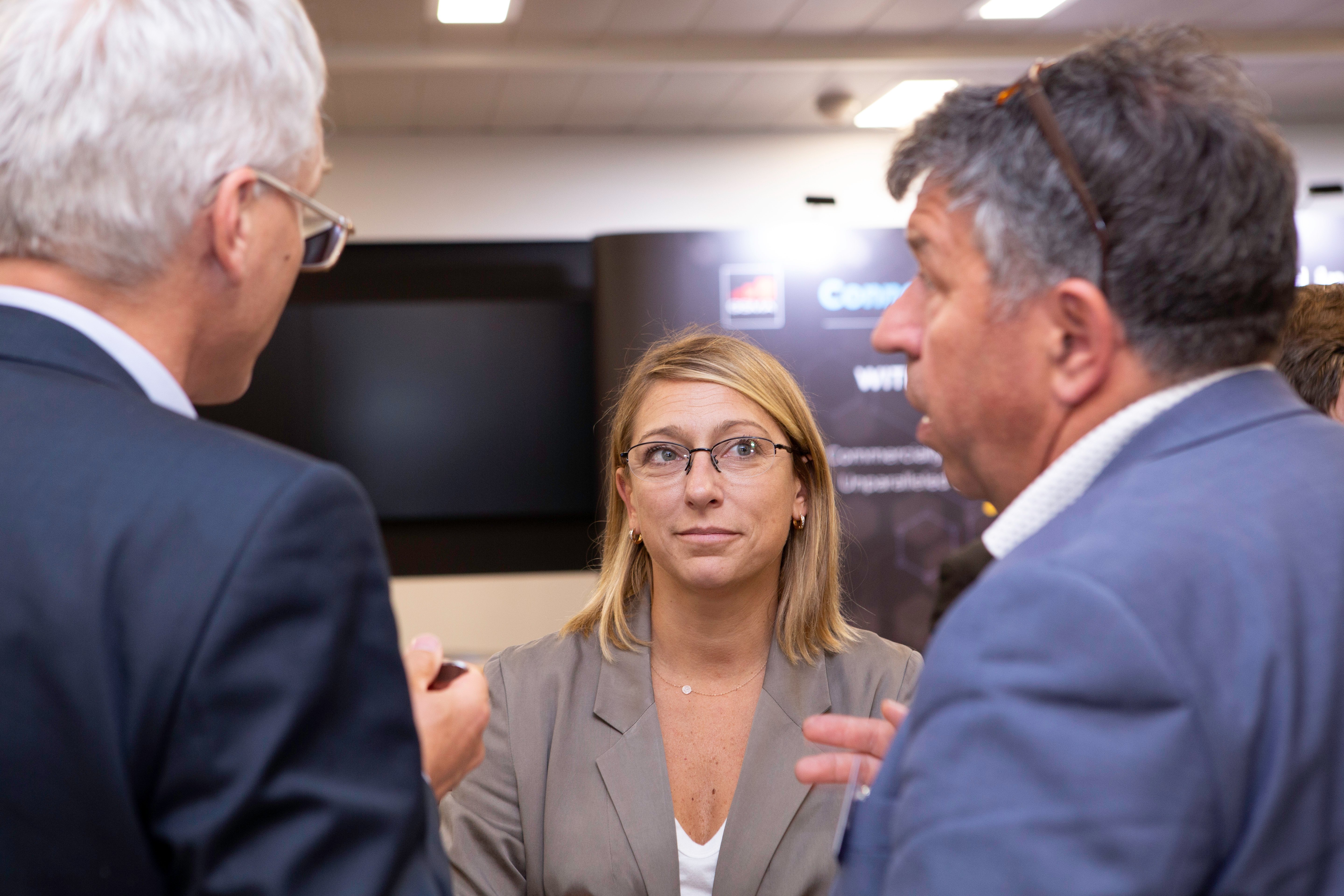 Three people in business attire discuss connected vehicles indoors. A blonde woman with glasses stands between two gray-haired men, one with glasses and the other with sunglasses on his head. A blurred display screen and lights are visible in the background.