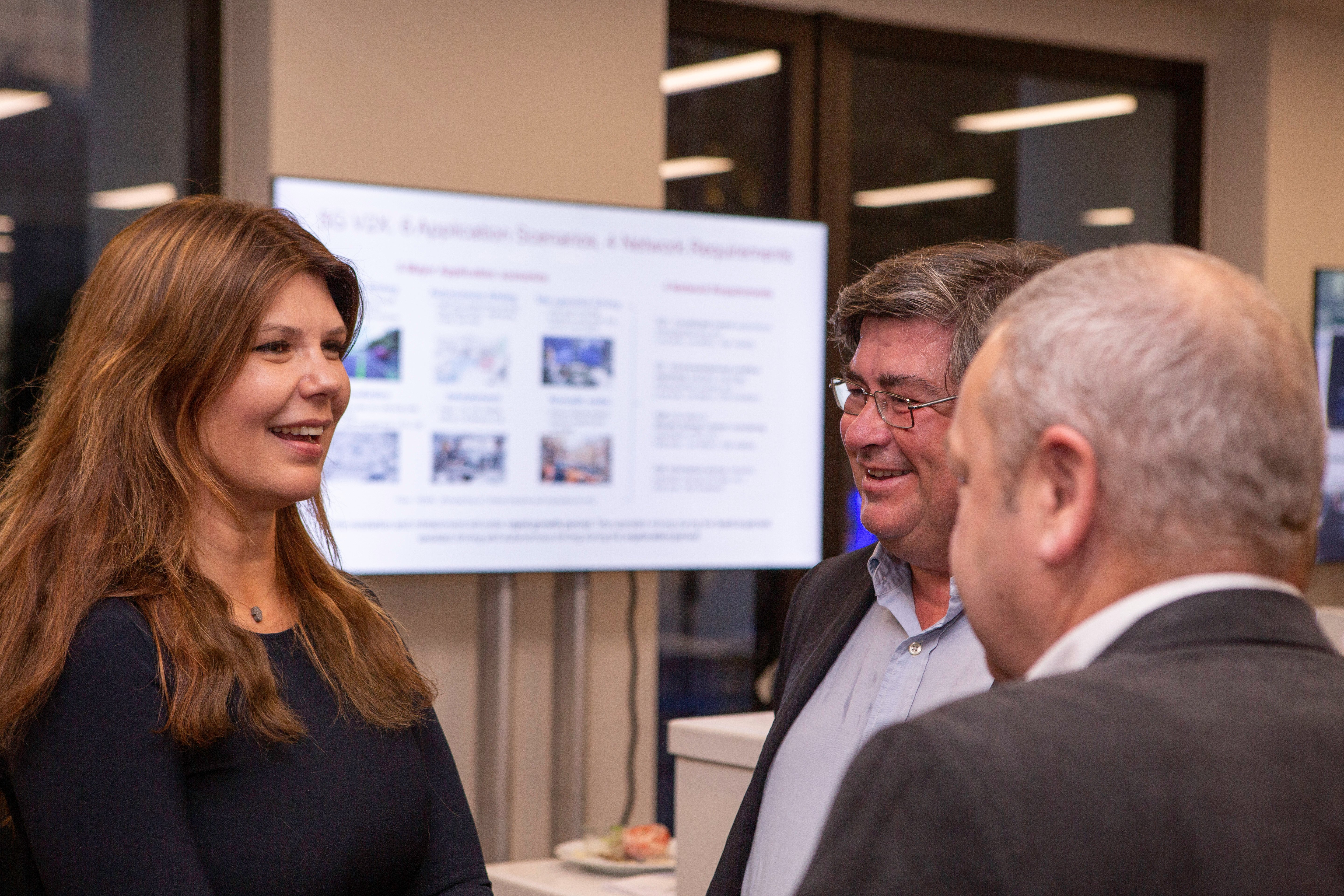 Three people, two men and one woman, stand indoors having a friendly conversation about connected vehicles. The woman, with long brown hair, smiles while listening. A blurred presentation screen and large windows are visible in the background. All are dressed in business attire.