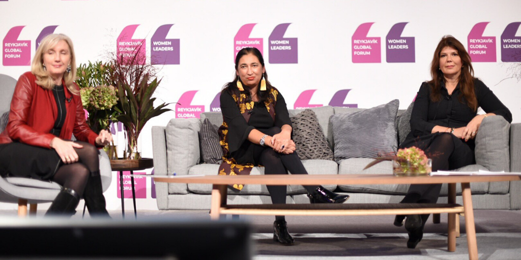 Three women sit on grey couches on a stage, engaged in a panel discussion. They wear black outfits, and one has a red jacket. The backdrop displays the "Reykjavik Global Forum Women Leaders" logo in purple and pink. Decorative plants and cushions are visible.