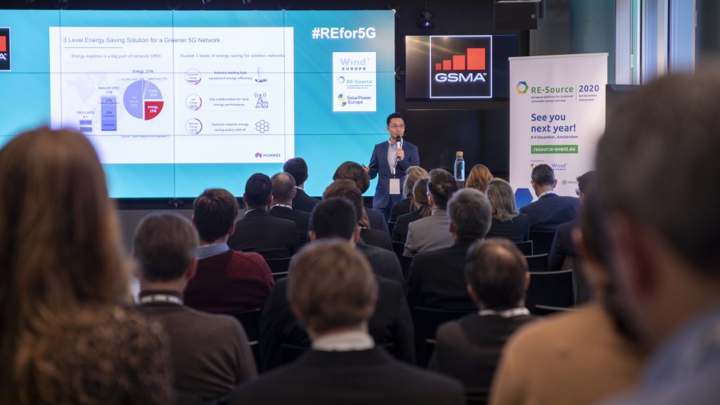 A man in a suit speaks on stage to an audience at a conference. Behind him, large screens display charts, logos, and presentation information related to energy savings for 5G. Signs for GSMA and “RE-Source 2020” are visible in the background.