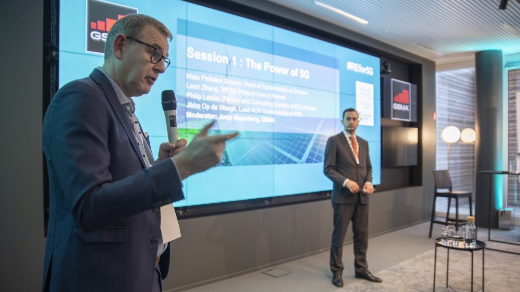 Two men in suits stand on a stage during a conference. One speaks into a microphone, gesturing, while the other stands nearby. Behind them, a large screen displays “Session 1: The Power of 5G” and details of the session and participants.