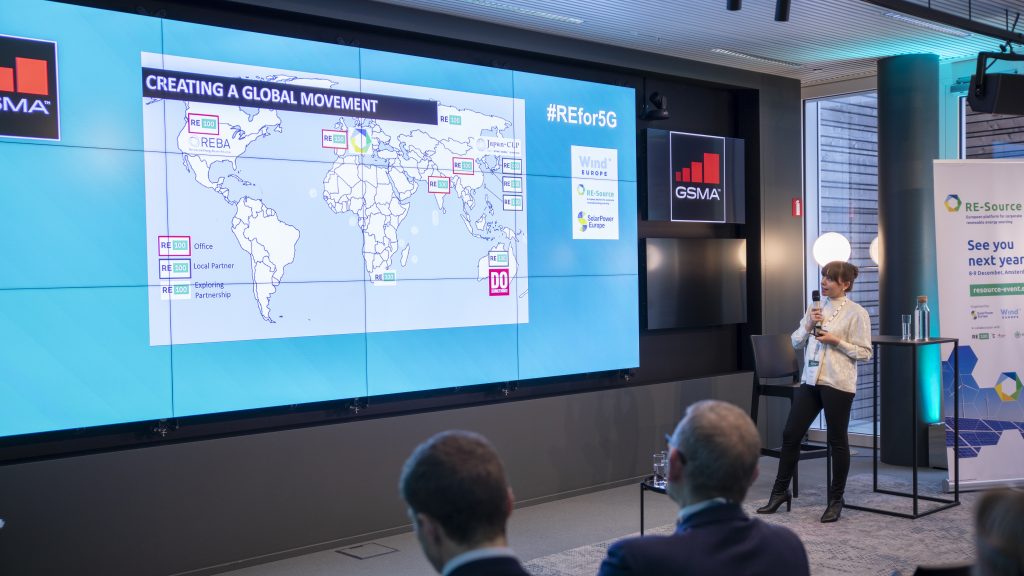 A woman stands with a microphone in front of a large screen displaying a world map labeled “Creating a Global Movement.” Several regions are highlighted. Seated audience members face her. A banner to the right reads “Re-Source, See you next year!” and windows line the background.