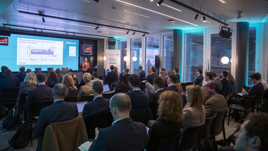 A large group of people sit facing a stage where a woman speaks at a podium. Multiple screens display graphs and text. The modern conference room has floor-to-ceiling windows and blue accent lighting. Attendees are engaged, some taking notes.