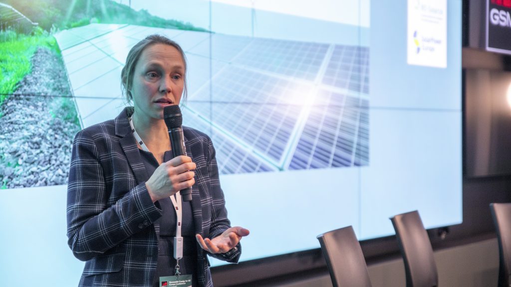 A woman in a plaid blazer speaks into a microphone, gesturing with her hand. She stands in front of a screen displaying an image of solar panels in a field. Empty chairs are visible beside her, suggesting a presentation or conference setting.