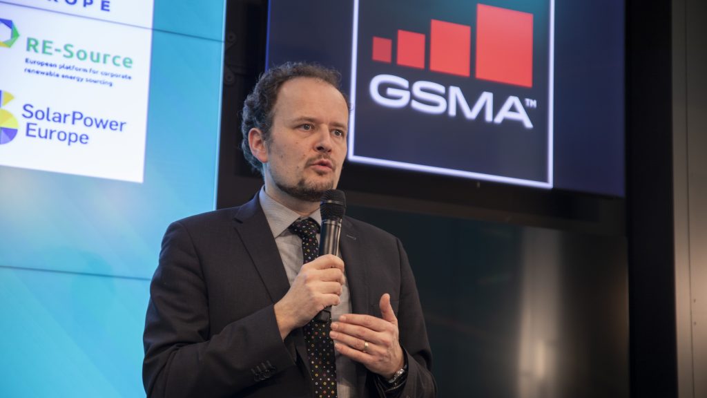 A man in a dark suit and tie speaks into a microphone. Behind him are two screens: one displays the GSMA logo with a red bar chart, and the other shows logos for RE-Source and SolarPower Europe. The setting appears to be a professional conference or event.