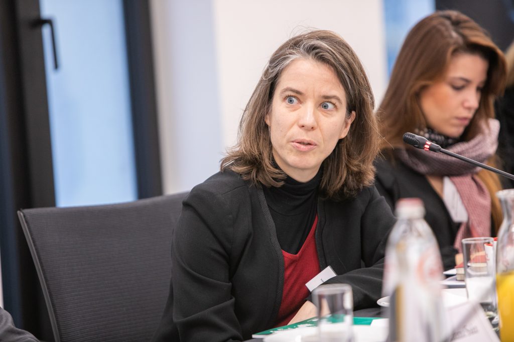 A woman with shoulder-length brown hair and a black blazer sits at a conference table, speaking or listening intently. A microphone is in front of her. Another woman sits nearby, focused on her notes. Bottles and papers are visible on the table.