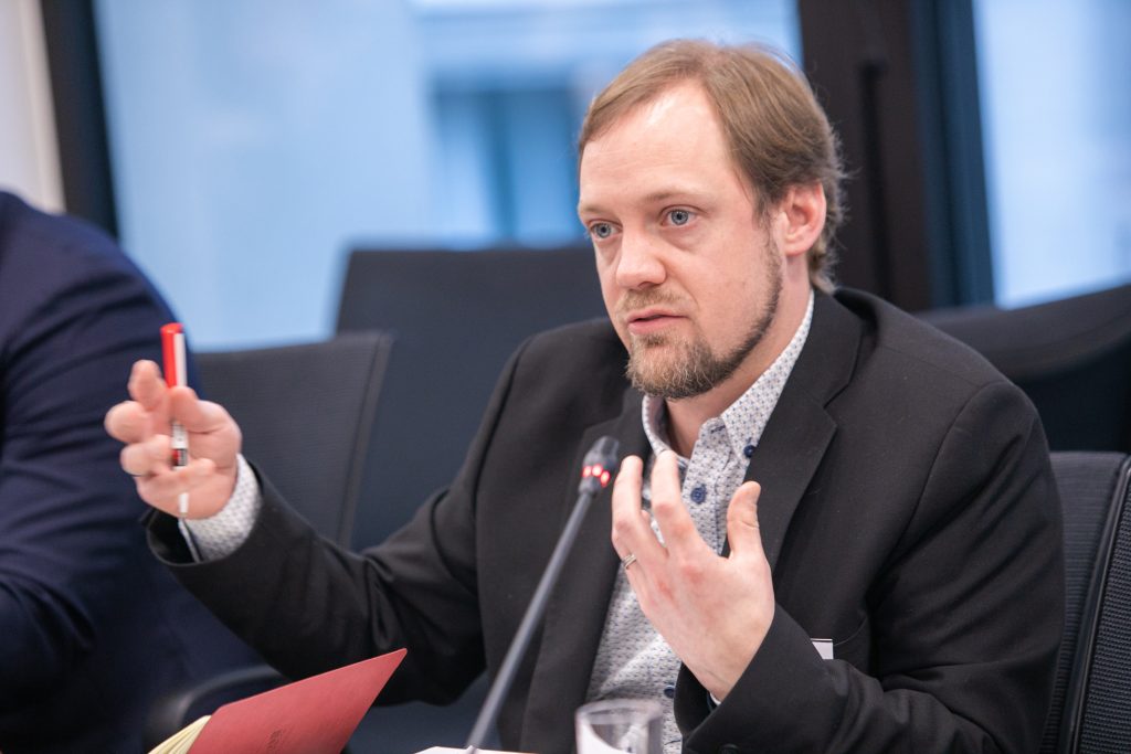 A man with light brown hair and a short beard speaks animatedly at a meeting. He wears a black blazer over a patterned shirt and gestures with one hand while holding a red pen in the other. A microphone and papers are visible in front of him.