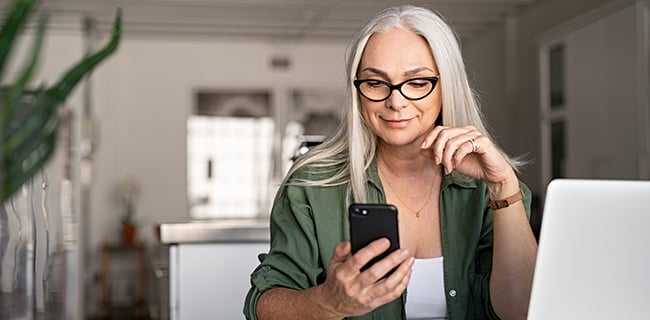 A smiling older woman with long gray hair and glasses sits at a table with a laptop, wearing a green shirt over a white top. She looks at her smartphone, appearing content, in a bright room with blurred modern decor in the background.