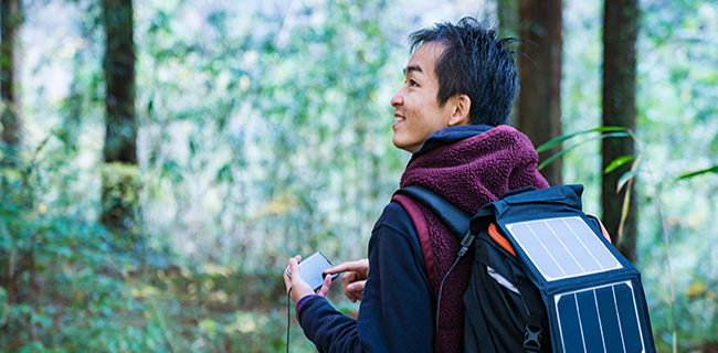 A person with short dark hair, wearing a dark jacket and maroon scarf, stands in a forest holding a smartphone. Their backpack has a visible solar panel on it. Sunlight filters through the tall trees and green foliage in the background.