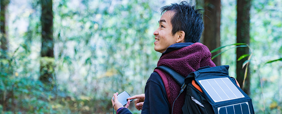 A person outdoors in a forest, smiling and looking to the side, holds a smartphone. They wear a burgundy jacket and carry a black backpack equipped with a solar panel. Green foliage and sunlight fill the background, creating a peaceful, natural atmosphere.