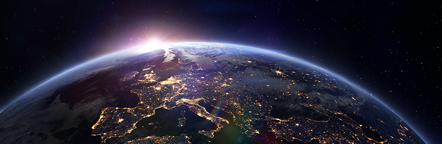 View of Earth from space showing sunrise at the horizon. City lights are visible across Europe and parts of Africa and Asia, with a curved edge of the planet fading into the darkness of space filled with scattered stars.