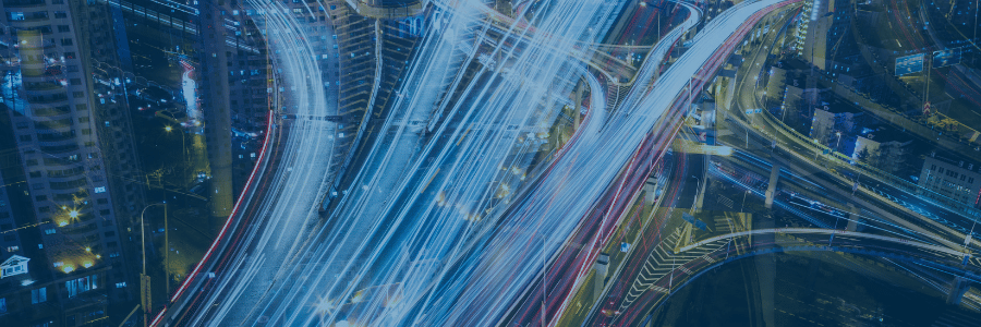 A night cityscape showing a complex network of highways and overpasses. Long exposure creates vivid light trails from vehicles, forming bright lines of blue, white, and red, emphasizing the movement and fast pace of urban traffic.