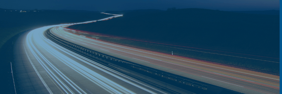 A long-exposure photo of a highway at night shows white and red light trails from vehicles, curving into the distance. The landscape is dark, with silhouettes of hills under a deep blue sky. No cars are visible, only their light trails.